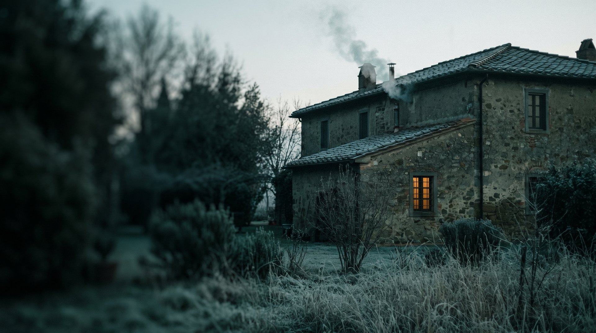 Casa in pietra all’alba d’inverno, con brina sul prato e il camino acceso